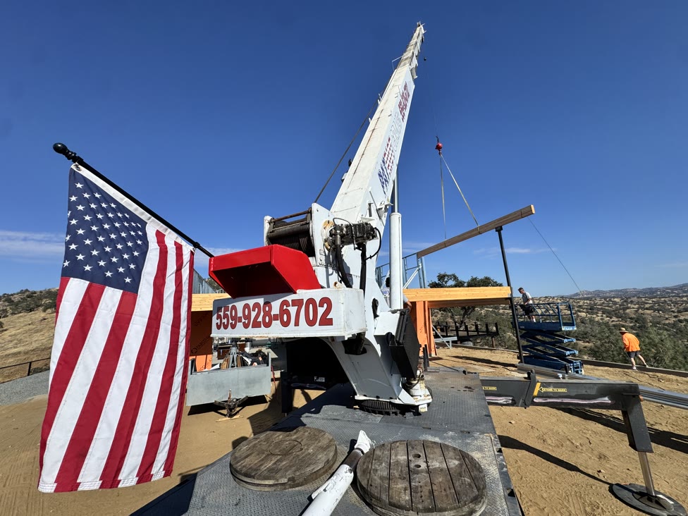 Patriot Crane lifting a glulam beam on a Fresno-area hilltop build, American flag on the boom, 559-928-6702 on the rig